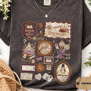 Może przedstawiać: Ciemnoszara koszulka z motywem książkowym, zawierająca napis "Chronicles of Discovery", lupę, sowę i inne elementy literackie. Widoczny jest złoty naszyjnik.