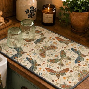 May include: A beige kitchen mat with a butterfly pattern, featuring orange, blue, and green butterflies. A matching dish towel hangs near a white sink. Two clear glasses sit on the mat, near a lit candle and a potted plant.