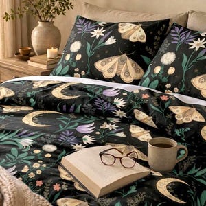 May include: A black duvet cover and pillowcases featuring a repeating pattern of beige moths, golden crescent moons, and various green, white, and purple wildflowers. An open book with brown-rimmed glasses and a beige mug sit on the bed.