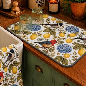 Puede incluir: Escena de cocina con un escurreplatos estampado y un paño de cocina a juego. El diseño incluye pájaros, limones e hortensias azules sobre un fondo blanco. La alfombrilla tiene un borde gris. También se ven un fregadero blanco y armarios verdes.