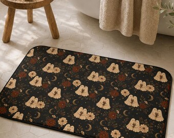 May include: A rectangular bath mat featuring a dark black background with a repeating pattern of small white ghosts holding flowers, interspersed with orange and red floral elements, green leaves, and small crescent moon shapes.