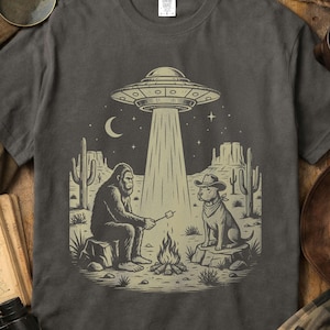 Pode incluir: Camiseta cinza escuro com uma cena desértica com um OVNI, um Bigfoot assando um marshmallow e um cachorro com chapéu de cowboy. O design inclui cactos, uma fogueira e um céu estrelado.