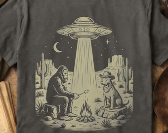 Puede incluir: Una camiseta gris carbón con un estampado gráfico de un Bigfoot y un perro con sombrero de vaquero sentados junto a una fogata en un desierto. Arriba, un platillo volador emite luz. El diseño está realizado en tinta de color beige claro.