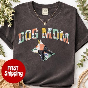 Puede incluir: Camiseta gris oscuro con las palabras "DOG MOM" en un diseño floral colorido. Una silueta de perro con un lazo negro está debajo del texto. La camiseta tiene mangas cortas y cuello redondo.
