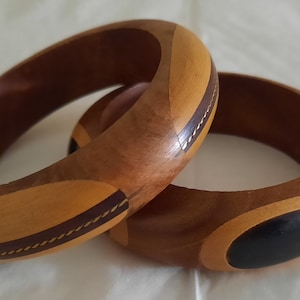 May include: Two wooden bangles with a brown, black, and yellow geometric pattern. The bangles have a thin gold line around the edges.
