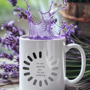 May include: White ceramic mug with a splash of purple liquid, featuring the text "I'm not ignoring you... I'm just buffering." The mug has a black and gray loading circle design. Lavender sprigs and flowers are in the background.
