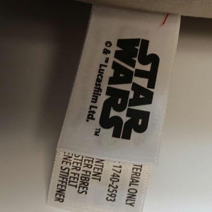 May include: A white tag with the Star Wars logo in black. The tag also includes the text "&copy; & &trade; Lucasfilm Ltd.", "MATERIAL ONLY", and "1740-2593". Additional text includes "NTENT", "TER FIBRES", "STER FELT", and "ENE STIFFENER".