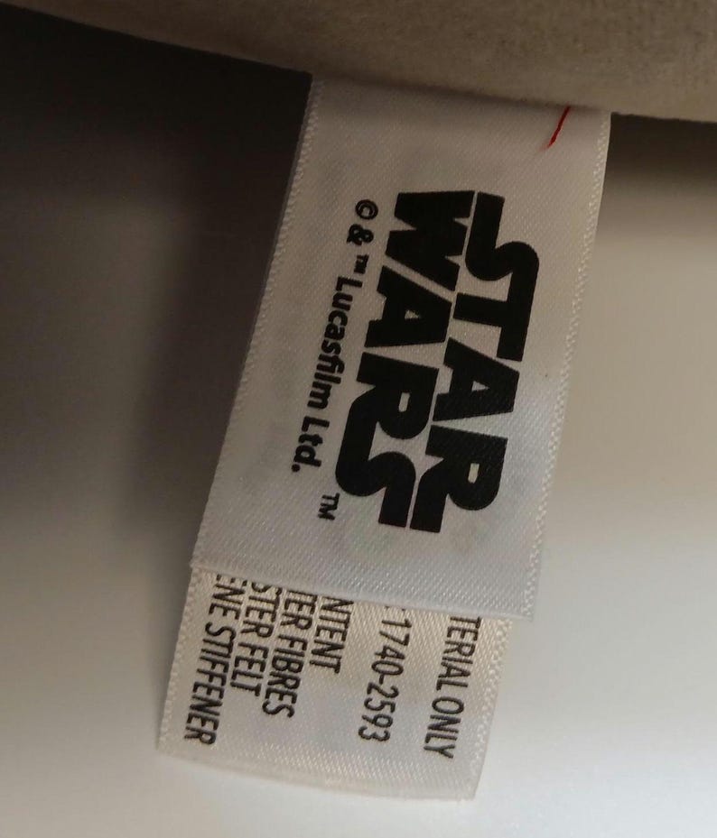 May include: A white tag with the Star Wars logo in black. The tag also includes the text "&copy; & &trade; Lucasfilm Ltd.", "MATERIAL ONLY", and "1740-2593". Additional text includes "NTENT", "TER FIBRES", "STER FELT", and "ENE STIFFENER".