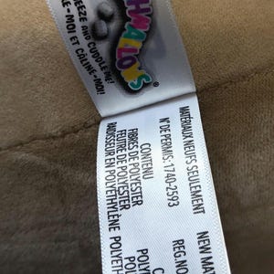 May include: A tag from a Squishmallows plush toy, featuring the brand name in colorful lettering. The tag includes the phrases "Squeeze and Cuddle Me!" and "New Material Only."