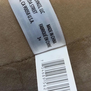 May include: A white tag with black text, including the company name KELLY TOYS HOLDINGS, LLC, address, and "MADE IN CHINA". The tag also includes a barcode and the text "3+". The tag is on a tan surface.