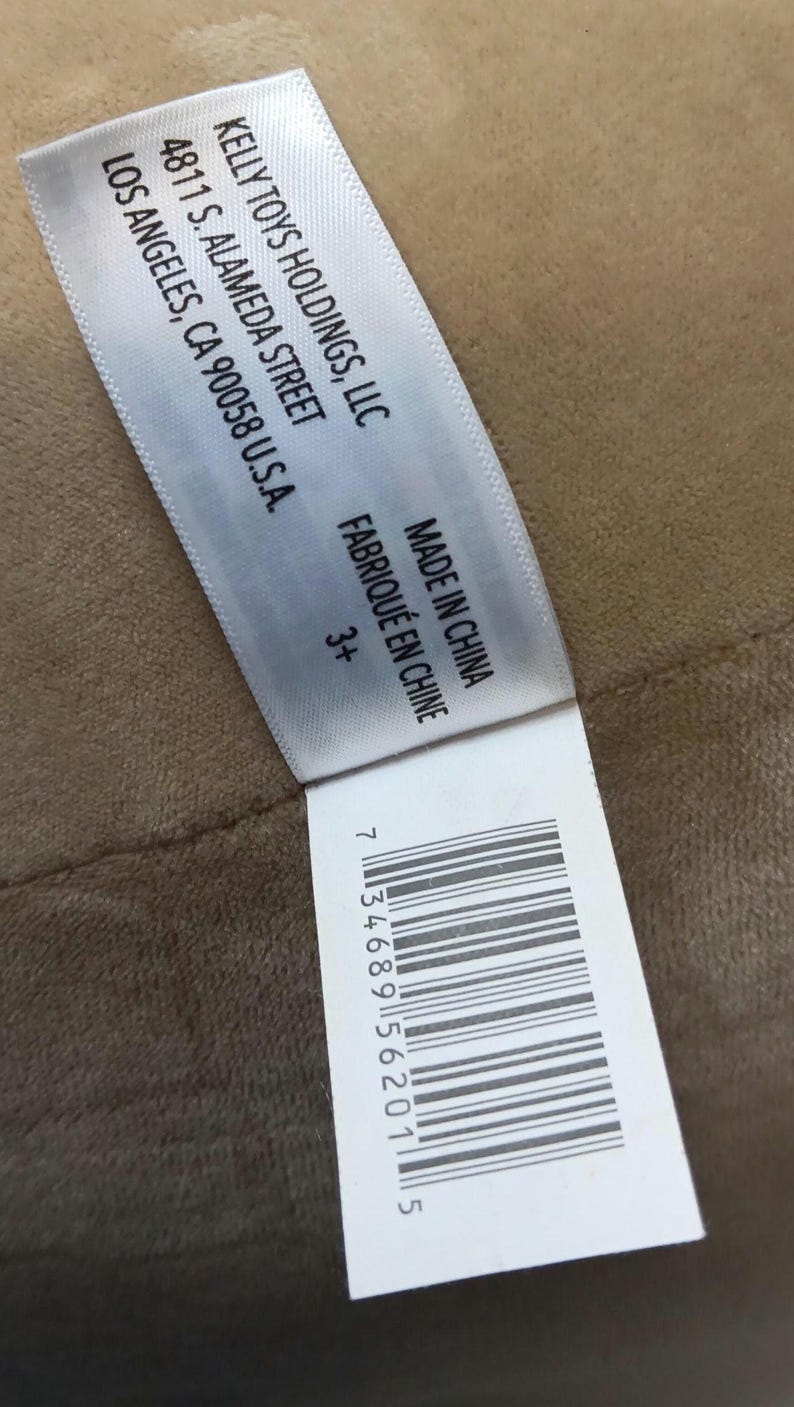 May include: A white tag with black text, including the company name KELLY TOYS HOLDINGS, LLC, address, and "MADE IN CHINA". The tag also includes a barcode and the text "3+". The tag is on a tan surface.