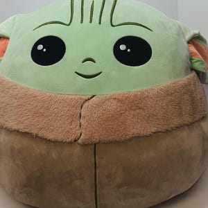 May include: A large plush toy shaped like a character from a popular science fiction series. The toy features a light green face with large black eyes, a small nose, and a stitched smile. It has a tan fur-like collar and a brown body.