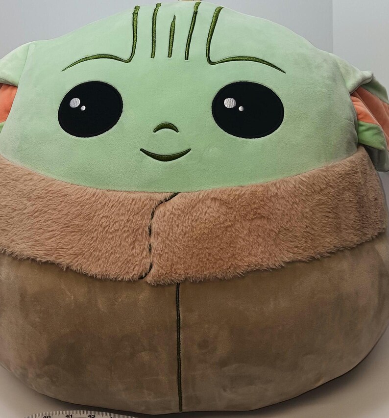 May include: A large plush toy shaped like a character from a popular science fiction series. The toy features a light green face with large black eyes, a small nose, and a stitched smile. It has a tan fur-like collar and a brown body.