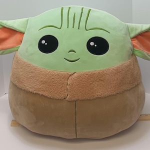 May include: A large plush toy shaped like a character with large black eyes, green skin, and large ears with orange insides. The toy has a tan-colored body and a brown furry collar. The toy is approximately 18 inches tall.