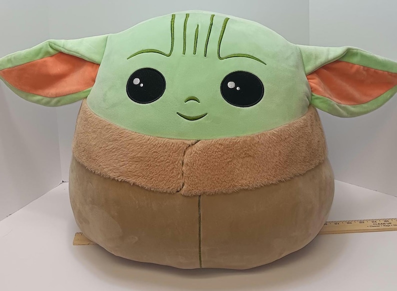 May include: A large plush toy shaped like a character with large black eyes, green skin, and large ears with orange insides. The toy has a tan-colored body and a brown furry collar. The toy is approximately 18 inches tall.