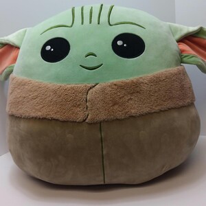 May include: A plush, round, green character with large black eyes, a small smile, and large ears with orange insides. The character has a tan furry collar and a brown body. The toy is a soft, cuddly pillow.