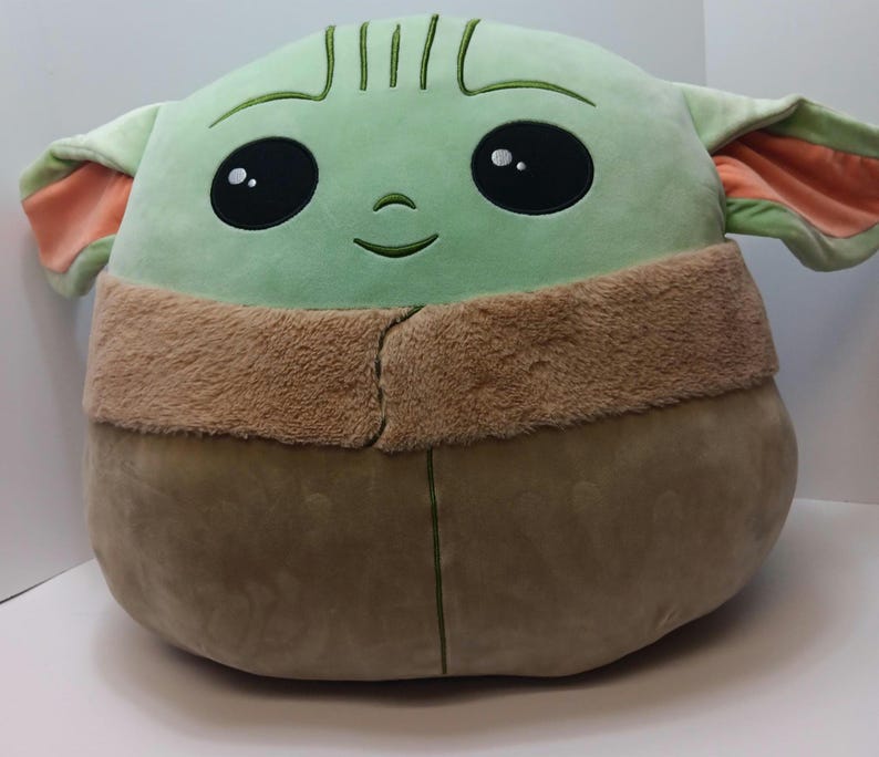 May include: A plush, round, green character with large black eyes, a small smile, and large ears with orange insides. The character has a tan furry collar and a brown body. The toy is a soft, cuddly pillow.