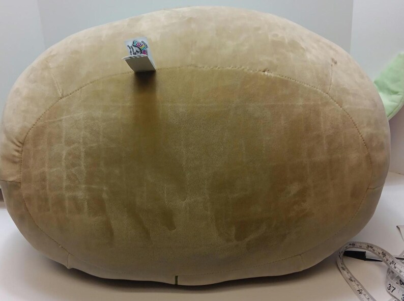 May include: A large, round, light brown plush pillow with a textured surface. A small tag is attached to the top. The pillow is likely for home decor or comfort.