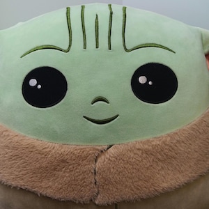 May include: A large plush toy featuring a character with a light green face, large black eyes, and a small smile. The toy has a tan, furry collar and embroidered details. The overall design is soft and cuddly.