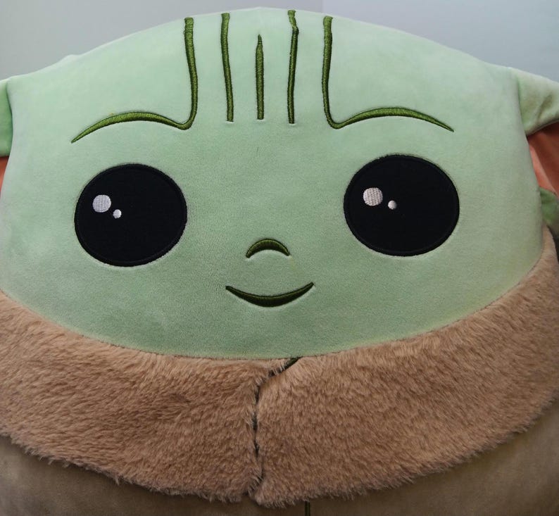 May include: A large plush toy featuring a character with a light green face, large black eyes, and a small smile. The toy has a tan, furry collar and embroidered details. The overall design is soft and cuddly.