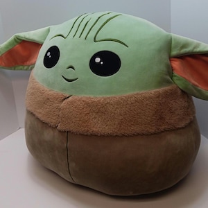 May include: A large plush toy shaped like a character with a large head, big black eyes, and large ears. The toy is primarily light green with brown and orange accents. The character has a stitched smile and embroidered details.