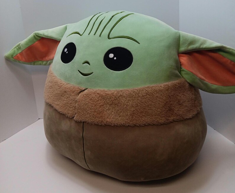 May include: A large plush toy shaped like a character with a large head, big black eyes, and large ears. The toy is primarily light green with brown and orange accents. The character has a stitched smile and embroidered details.
