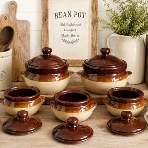 May include: A collection of brown and cream-colored bean pots with lids, displayed on a wooden surface. The pots vary in size and shape, with a two-tone design. A sign reads "BEAN POT" with text below. Wooden spoons and a cutting board are in the background.