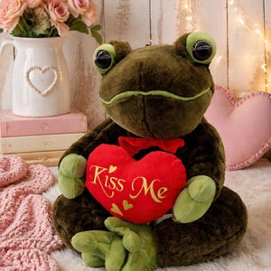 May include: A plush frog toy with large green eyes and green accents. The frog holds a red heart-shaped pillow with the words "Kiss Me" in gold script. The frog is brown and green, with a red bow.