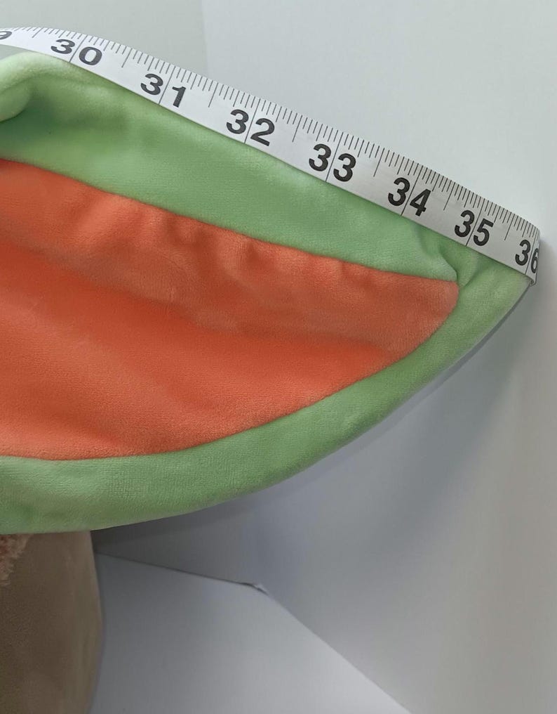 May include: A plush, green and orange costume ear with a measuring tape draped over it. The tape reads from 30 to 36 inches. The ear is green around the edges and orange in the center.