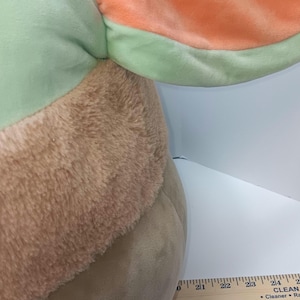 May include: A plush toy featuring a large head and ears. The ears are a light green with an orange interior. The head has a tan, furry texture and a smooth, light brown body. A wooden ruler is placed next to the toy.