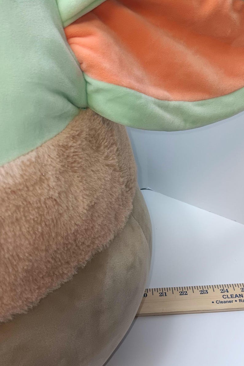 May include: A plush toy featuring a large head and ears. The ears are a light green with an orange interior. The head has a tan, furry texture and a smooth, light brown body. A wooden ruler is placed next to the toy.