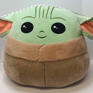 May include: A large plush toy shaped like a character with a light green face, large black eyes, and a small smile. The ears are light green with orange insides. The plush is tan and brown. A measuring tape is draped over the top of the plush.