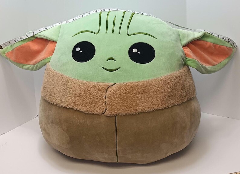 May include: A large plush toy shaped like a character with a light green face, large black eyes, and a small smile. The ears are light green with orange insides. The plush is tan and brown. A measuring tape is draped over the top of the plush.