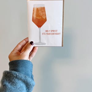 May include: A birthday card with an illustration of a glass of orange juice with ice cubes and a slice of orange. The text on the card reads "Holy Spritz! It's your birthday!"