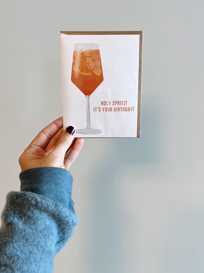 May include: A birthday card with an illustration of a glass of orange juice with ice cubes and a slice of orange. The text on the card reads "Holy Spritz! It's your birthday!"