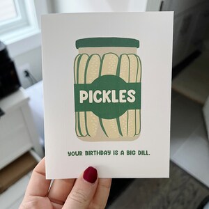 May include: A white card with a green jar of pickles. The jar has a green label that says 'PICKLES'. The text below the jar says 'YOUR BIRTHDAY IS A BIG DILL.'