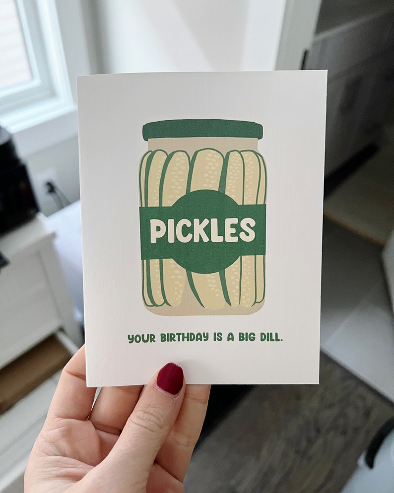 May include: A white card with a green jar of pickles. The jar has a green label that says 'PICKLES'. The text below the jar says 'YOUR BIRTHDAY IS A BIG DILL.'