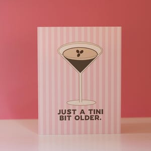 Op de afbeelding: Een wenskaart met een roze en witte streepjesachtergrond. Het heeft een martini-glas illustratie gevuld met een donkere vloeistof en drie koffiebonen. De tekst "JUST A TINI BIT OLDER." staat eronder.