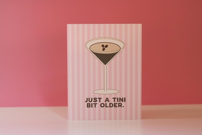 Op de afbeelding: Een wenskaart met een roze en witte streepjesachtergrond. Het heeft een martini-glas illustratie gevuld met een donkere vloeistof en drie koffiebonen. De tekst "JUST A TINI BIT OLDER." staat eronder.