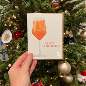 May include: A greeting card with an illustration of a glass of orange juice with a slice of orange in it. The text on the card reads "Holy Spritz! It's your birthday!"