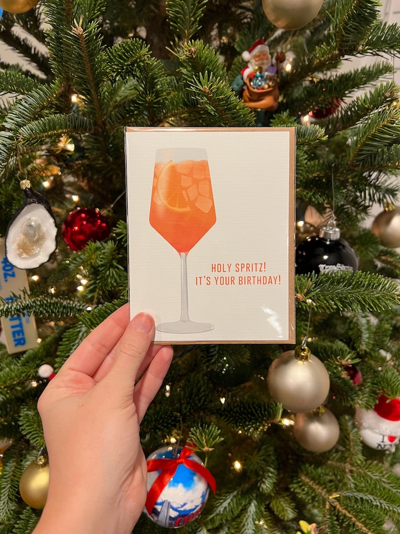 May include: A greeting card with an illustration of a glass of orange juice with a slice of orange in it. The text on the card reads "Holy Spritz! It's your birthday!"