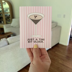 Op de afbeelding: Een wenskaart met een roze en witte streepjesachtergrond. De kaart toont een martini-glas illustratie met een donkerbruin drankje en twee koffiebonen. De tekst "JUST A TINI BIT OLDER" staat eronder.