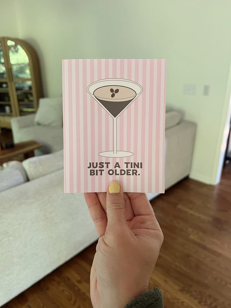 Op de afbeelding: Een wenskaart met een roze en witte streepjesachtergrond. De kaart toont een martini-glas illustratie met een donkerbruin drankje en twee koffiebonen. De tekst "JUST A TINI BIT OLDER" staat eronder.