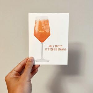 May include: A white card with an illustration of a glass of orange juice with ice and an orange slice. The text on the card reads "Holy Spritz! It's your birthday!"