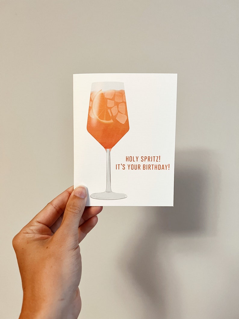 May include: A white card with an illustration of a glass of orange juice with ice and an orange slice. The text on the card reads "Holy Spritz! It's your birthday!"