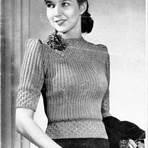 Puede incluir: Fotografía en blanco y negro de una mujer con un suéter de punto y un sombrero. El suéter tiene canalé vertical y mangas cortas con puños estampados. El sombrero es oscuro con un elemento decorativo. El texto "PATONS BEEHIVE FINGERING, 3-PLY" es visible.