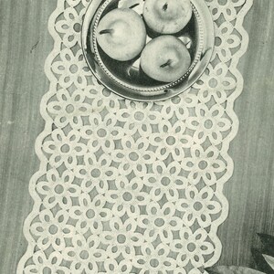 May include: A white crocheted table runner with a delicate floral pattern. The runner is laid on a wooden table with a silver bowl containing three apples.