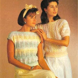 以下が含まれることがあります： 2人の女性が編み物の上着を着ています。左側の女性は、薄い青と白のストライプのトップに、黄色のリボンを髪に付けています。右側の女性は、白と薄いピンクの編み物の上着を着ています。2人とも笑顔です。
