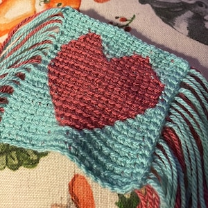 May include: A small woven heart-shaped wall hanging with a pink heart in the center and a light blue background. The hanging has long pink and blue fringe on the bottom.