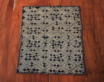 May include: A rectangular textile with a geometric pattern in black, tan, and white. The design features interlocking diamond shapes and a dark gray border. The textile is displayed on a wooden surface.